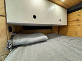 Cozy RV rental bed with overhead cabinets, a soundbar, and warm wood paneling, ready for a comfortable rest.