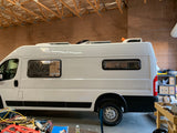 White RV rental camper van, showing custom windows & roof units, in a workshop with tools on the floor.