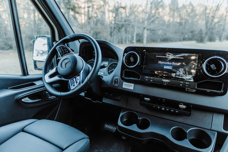 Inside a Mercedes-Benz RV rental: a clean, modern dashboard with steering wheel and large display. Road visible ahead.