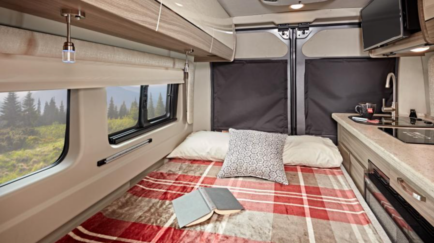 Cozy RV rental interior: bed with a plaid blanket, kitchen, and scenic window views of nature.