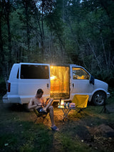 A man relaxes by his brightly lit **RV rental** van at a dark forest campsite. Inside, a cozy bed is visible.