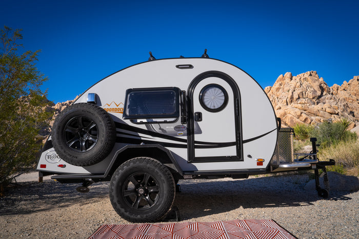 This compact teardrop RV rental, ideal for desert camping adventures, is parked on gravel with rocky terrain and a clear blue sky.