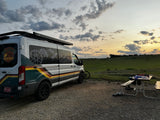 Camper van rental under a vibrant sunset with a custom-designed van, parked on gravel next to a picnic table in a grassy field.