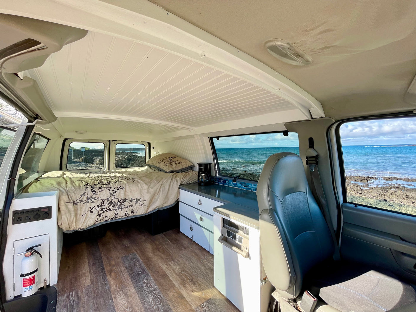 Cozy camper van interior with ocean view, perfect for an RV rental adventure. Features bed, kitchen, & scenic backdrop.