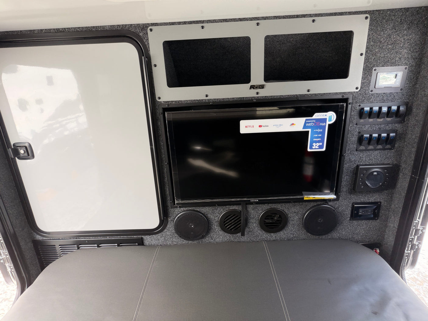 RV rental interior with a 32" Smart TV, JBL audio system, and a control panel for switches, above a gray bed area.