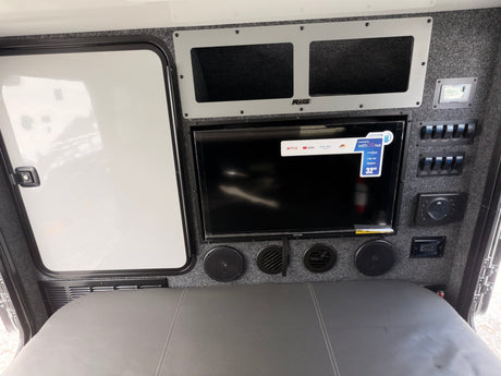 RV rental interior with a 32" Smart TV, JBL audio system, and a control panel for switches, above a gray bed area.