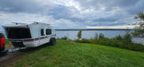 A white teardrop camper trailer (possibly an RV rental) sits on a grassy lakeside campsite under a cloudy sky.