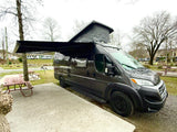 This dark gray camper van, possibly an RV rental, is set up at a campsite with its awning extended and pop-top raised.