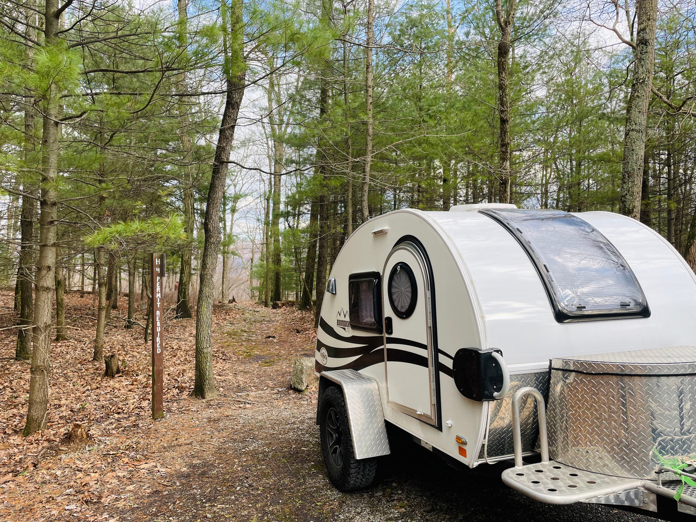 This white teardrop camper, an ideal RV rental, is parked at a quiet wooded campsite amidst trees and fallen leaves.