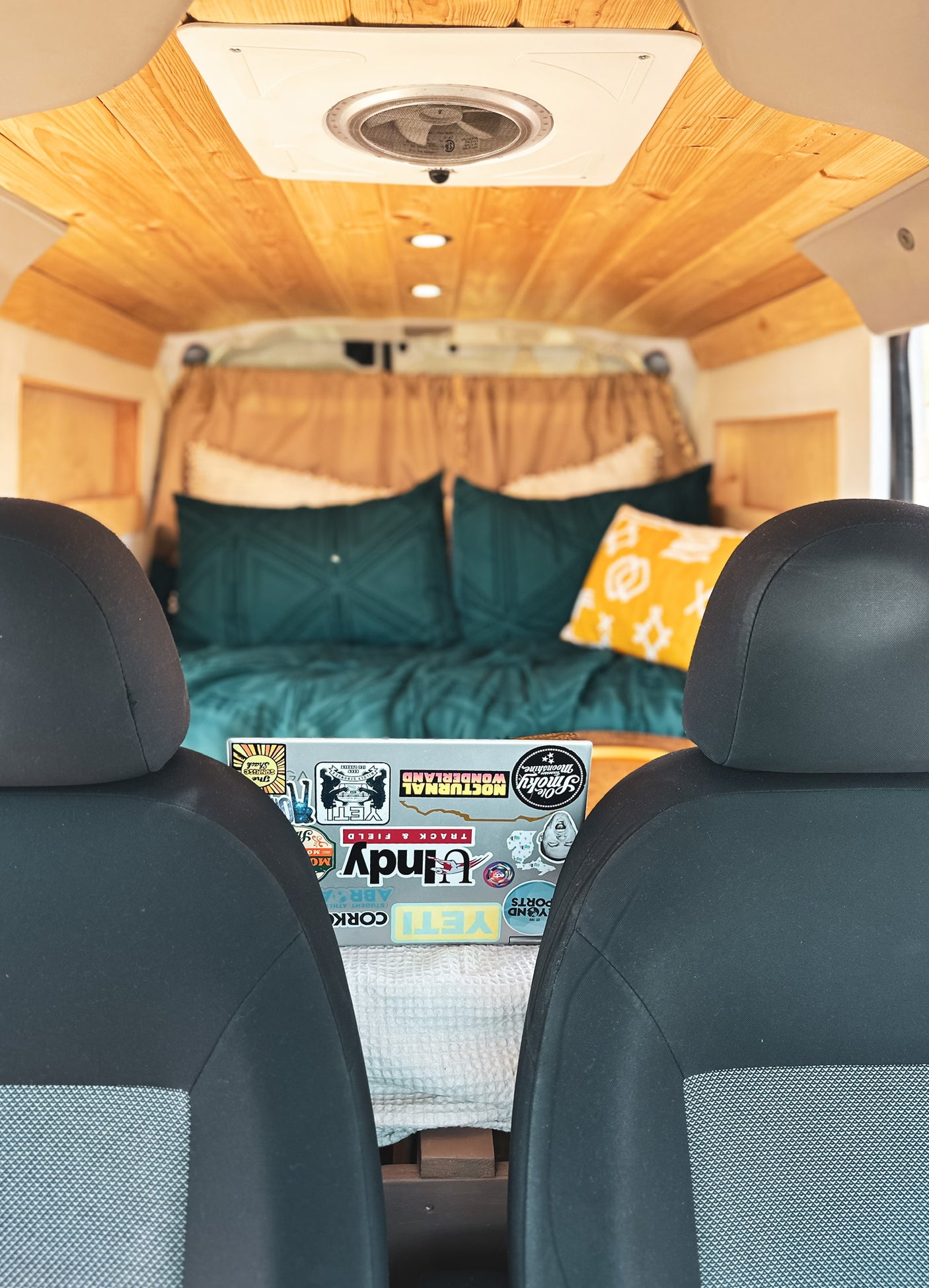 Cozy RV rental interior. View from front seats to bed with teal/yellow pillows. Wooden ceiling & laptop with stickers.