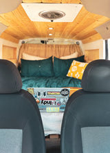 Cozy RV rental interior. View from front seats to bed with teal/yellow pillows. Wooden ceiling & laptop with stickers.