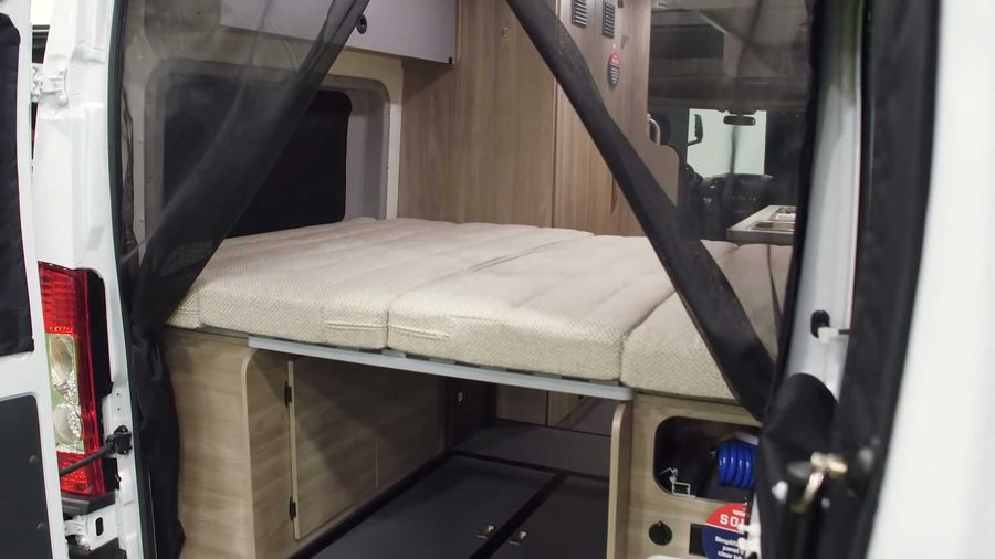 Interior of an RV rental van: convertible bed, under-bed storage, and a black mesh screen at the open rear doors.