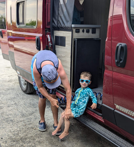 Dad uses RV rental outdoor shower on child's legs. Perfect for family adventures in a red Winnebago.