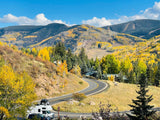 An RV rental navigates a winding mountain road, surrounded by vibrant fall foliage. Distant peaks reveal ski trails under a blue sky.
