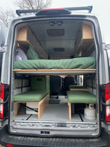Spacious RV rental camper van interior with custom wooden storage, a cozy bed, and green cushioned seating.