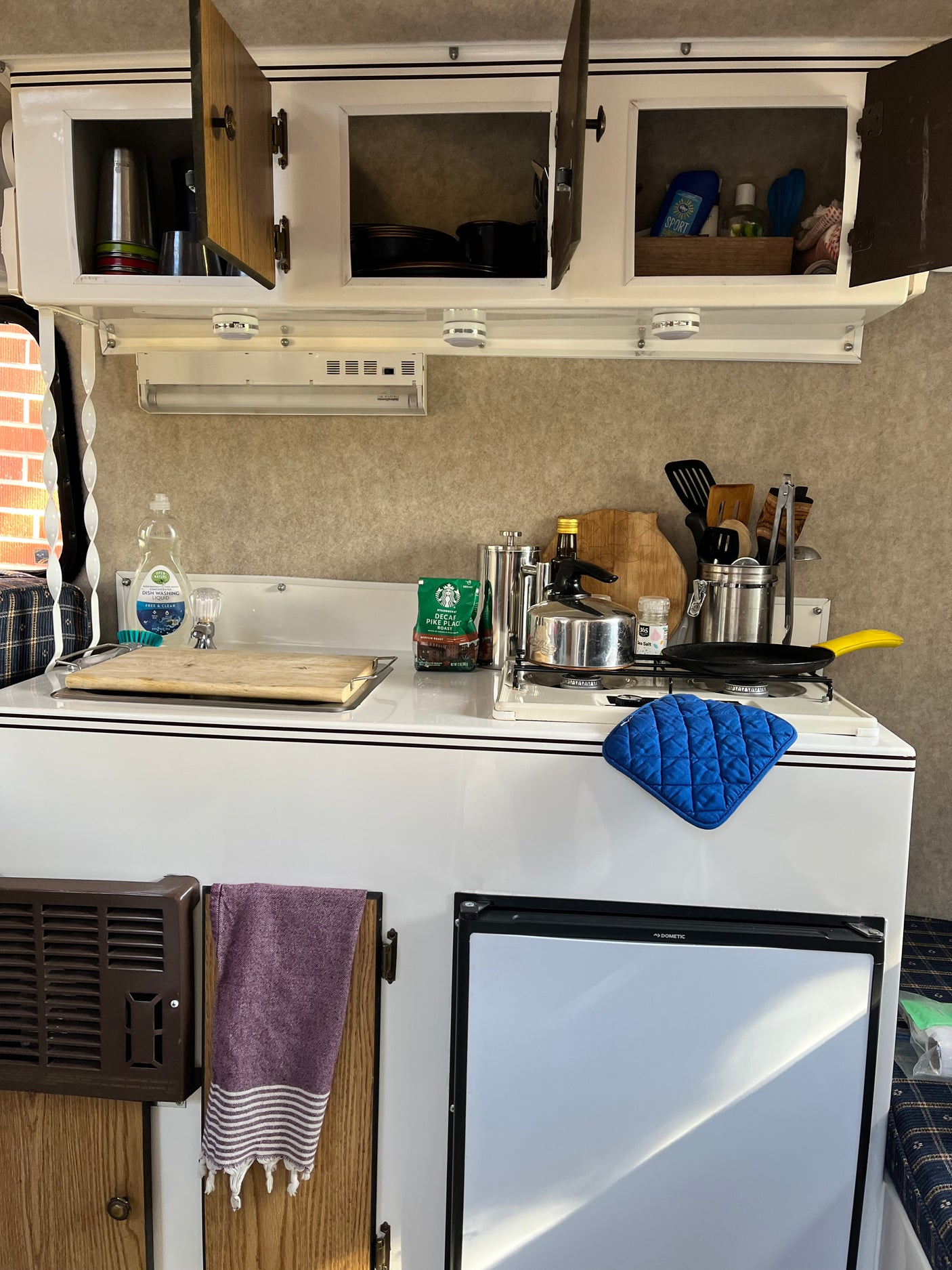 Compact RV rental kitchen with open cabinets, a stove, Dometic fridge, and cooking essentials ready.