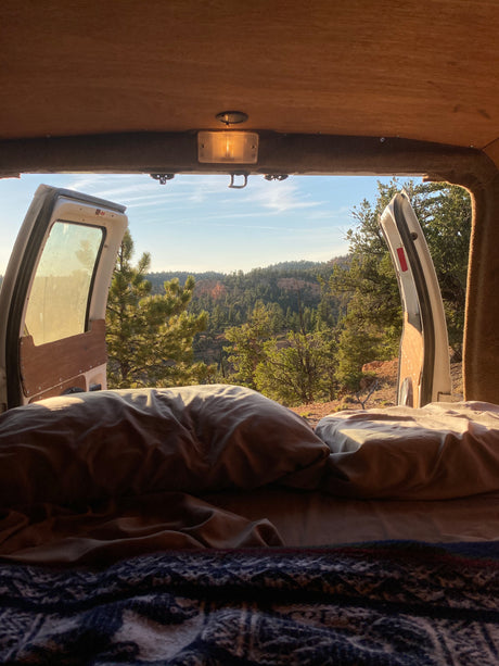 Inside a cozy **RV rental**, a bed faces open rear doors revealing a serene mountain forest view, perfect for **van life** adventures.