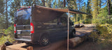 Dark grey Carado Banff RV rental with its awning extended, parked in a sunlit forest campsite.