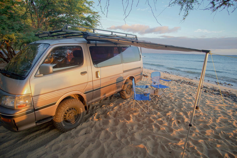 A converted campervan with an awning and chairs is perfectly set up on a sandy beach at sunset. Ideal for an RV rental adventure!|This RV rental campervan offers beachside comfort. Open door reveals bed — photo 1