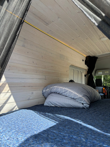Bright RV rental interior: wood-paneled walls, warm LED light, blue bed. Sunlit space for comfortable van life travel.