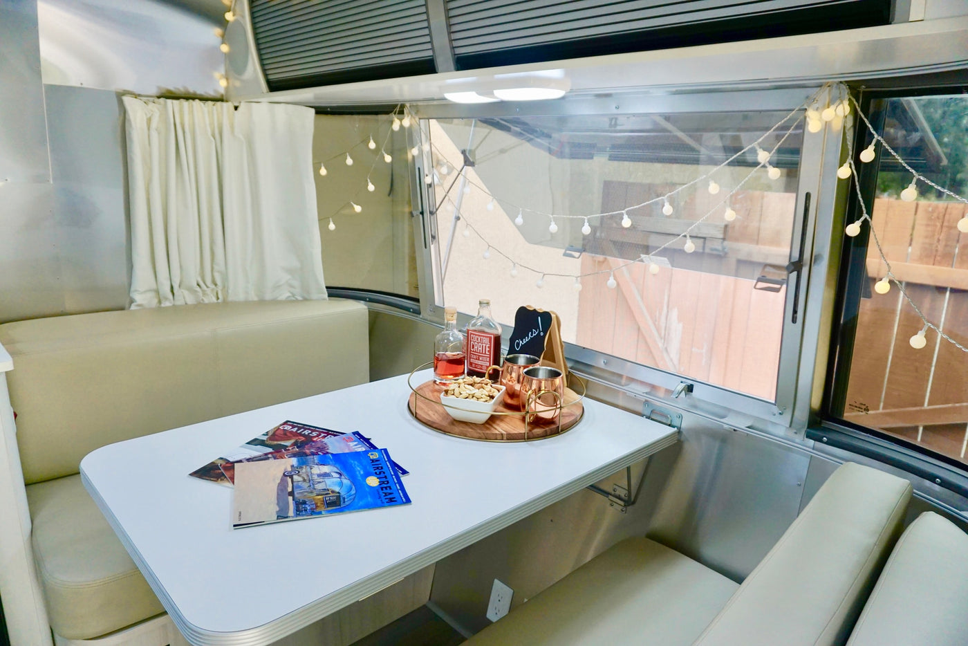 Cozy Airstream RV rental interior features a dining table with drinks, snacks, magazines, and charming string lights. — photo 8