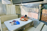 Cozy Airstream RV rental interior features a dining table with drinks, snacks, magazines, and charming string lights.