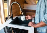 Inside an RV rental, a man fills a mug at the kitchen sink. Features a modern faucet, stove, and cozy wood interior.
