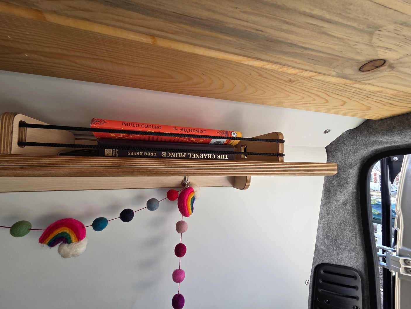 Cozy RV rental interior with a secure wooden bookshelf holding "The Alchemist" and "The Charnel Prince," plus felt rainbow decor.