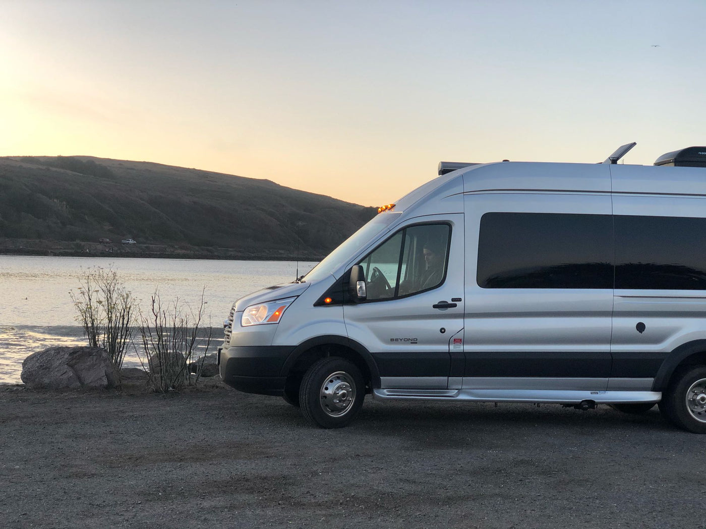  and two French Bulldogs by a tranquil lake surrounded by bare trees. Ideal for travel.|Experience adventure! Silver RV rental parked near a majestic red rock cliff. People explore the scenic landscape under blue skies.|Silver RV rental camper van parked at waterside — photo 3