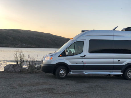 Silver RV rental camper van parked at waterside, bathed in sunset hues, ready for adventure.