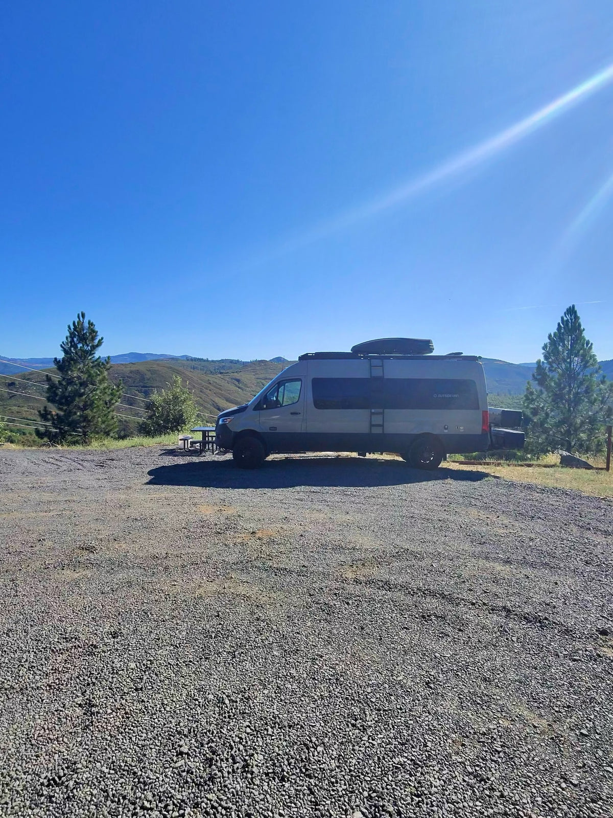 **RV rental** parked on a sunny gravel lot with mountain views & pine trees. Ideal for a serene outdoor escape.|This **rv rental** camper van is ready for adventure among towering redwood trees with a person and dog on a dirt road.|An RV rental camper van with a hitched SUV parked on a gravel path