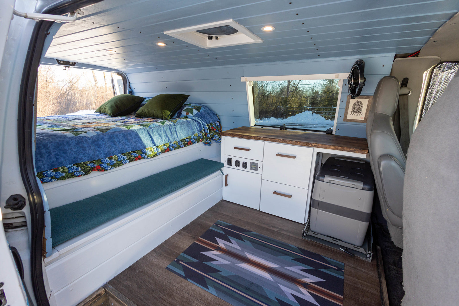 This cozy rv rental van's interior features a bed, kitchenette, and fridge on a slide-out, with light blue walls and smart storage.