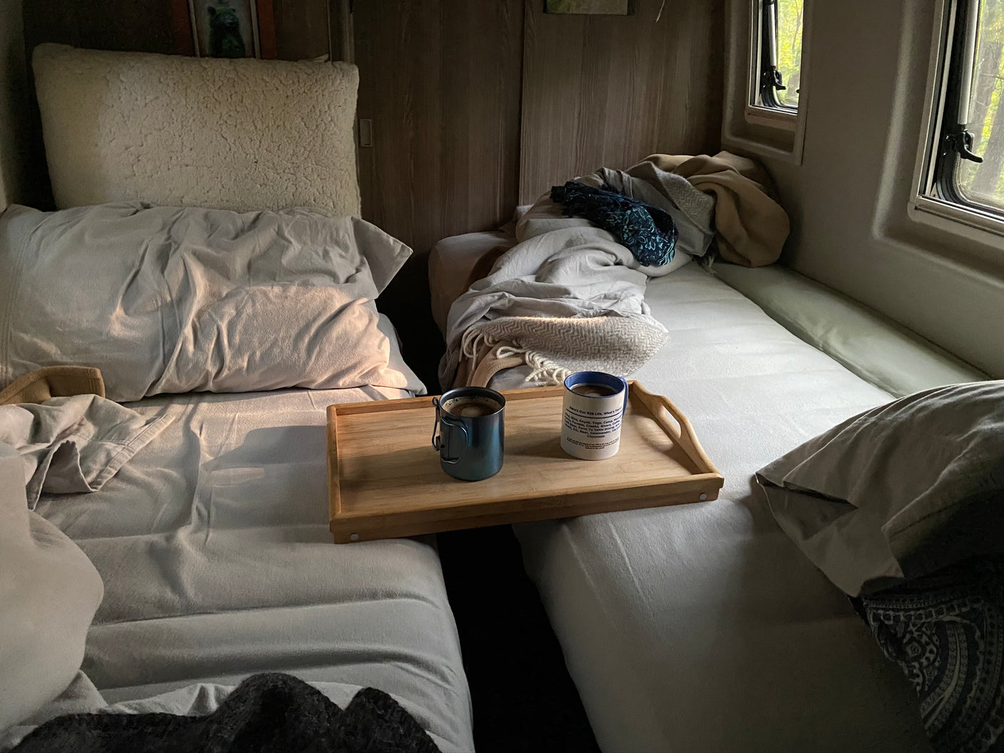 Cozy rv rental interior with two beds, featuring a wooden tray with coffee mugs and windows showing trees outside.