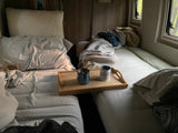 Cozy rv rental interior with two beds, featuring a wooden tray with coffee mugs and windows showing trees outside.