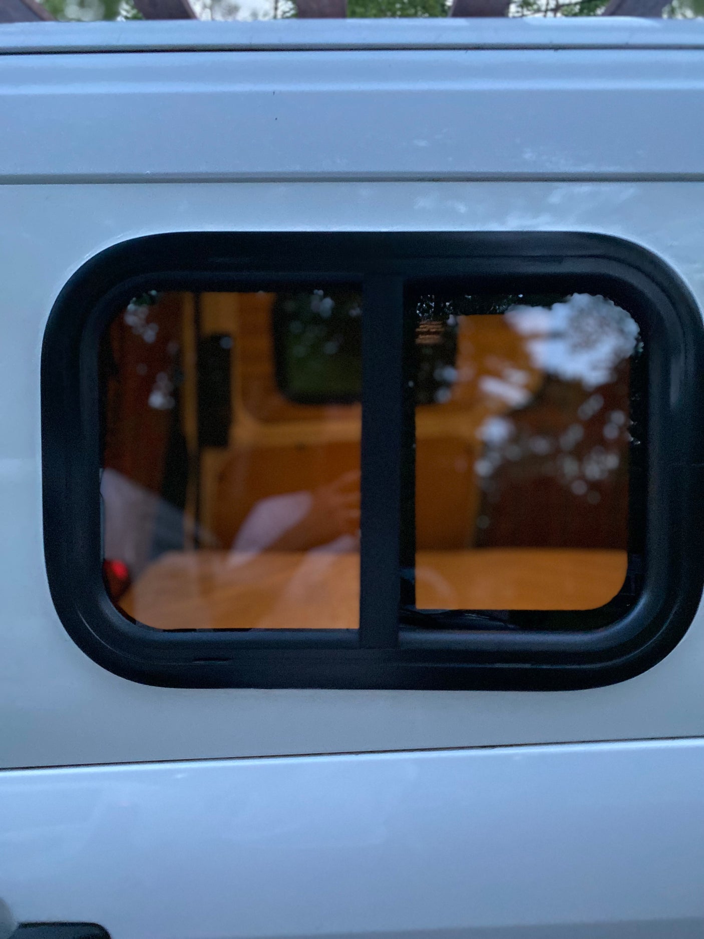 Side view of a white rv rental van's window. The dark-tinted window has a black frame, showing interior wood and blurred reflections.
