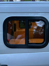 Side view of a white rv rental van's window. The dark-tinted window has a black frame, showing interior wood and blurred reflections.