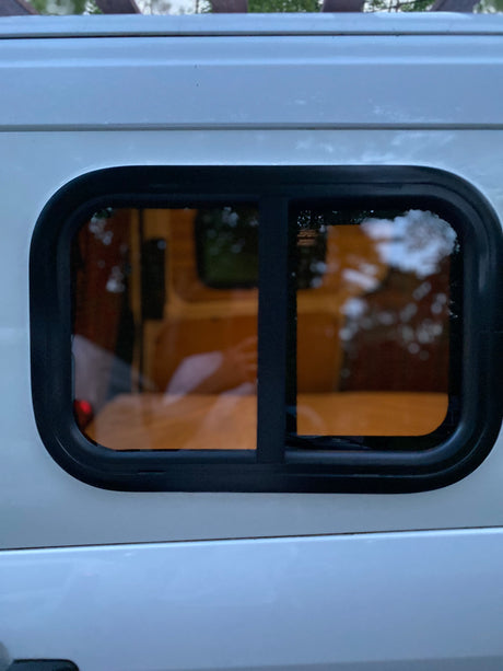 Side view of a white rv rental van's window. The dark-tinted window has a black frame, showing interior wood and blurred reflections.