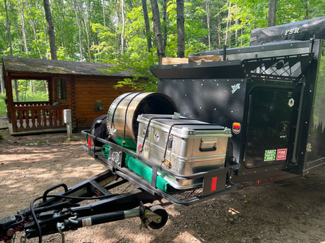 This RV rental Boreas camper trailer, loaded with gear, is set up at a wooded campsite beside a log cabin.