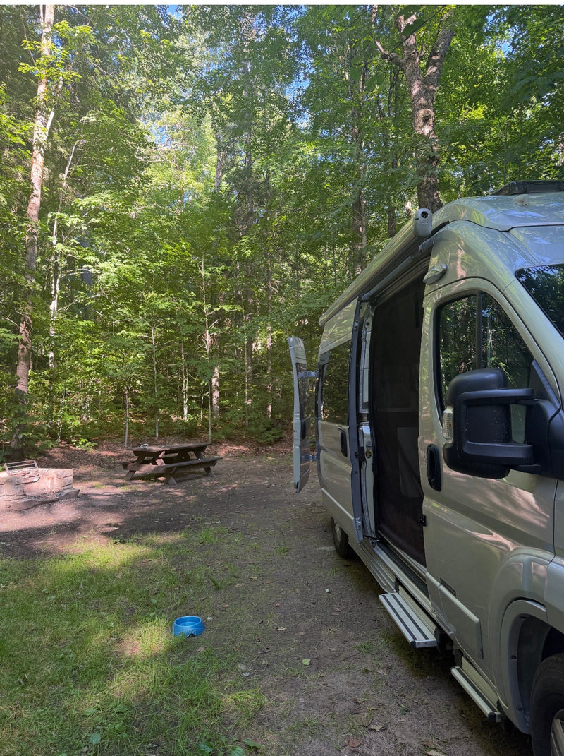 RV rental parked at a shaded, forested campsite with an open door, a fire pit, picnic table, and pet bowl. — photo 15