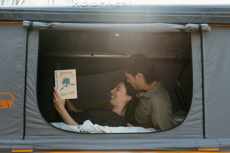 Cozy 'rv rental' rooftop tent: A smiling couple relaxes, reading "Travels in Alaska" inside their comfortable Roofnest.