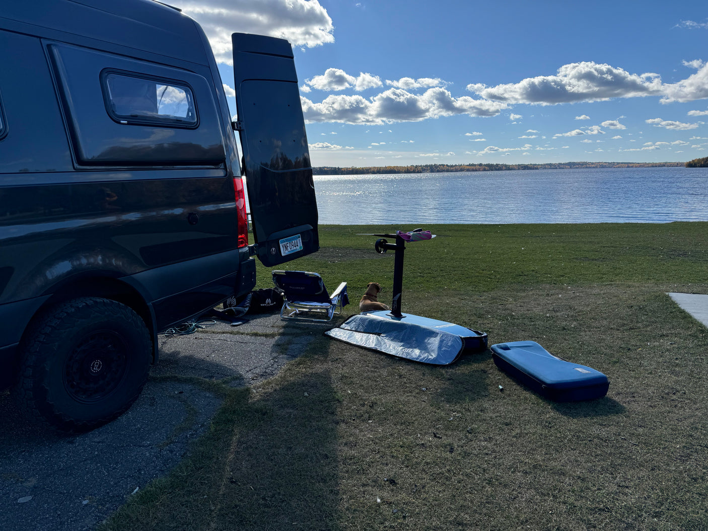 A dark camper van by a lake with an e-foil board and a dog. Ideal rv rental for hydrofoiling adventures.
