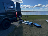 A dark camper van by a lake with an e-foil board and a dog. Ideal rv rental for hydrofoiling adventures.