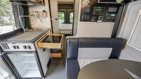 This RV rental interior shows a compact kitchen with sink, fridge, pull-out cooktop, and a dinette leading to a rear bedroom.