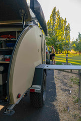 RV rental setup! Rear hatch of a beige teardrop camper open for kitchen access, table extended. Person nearby.