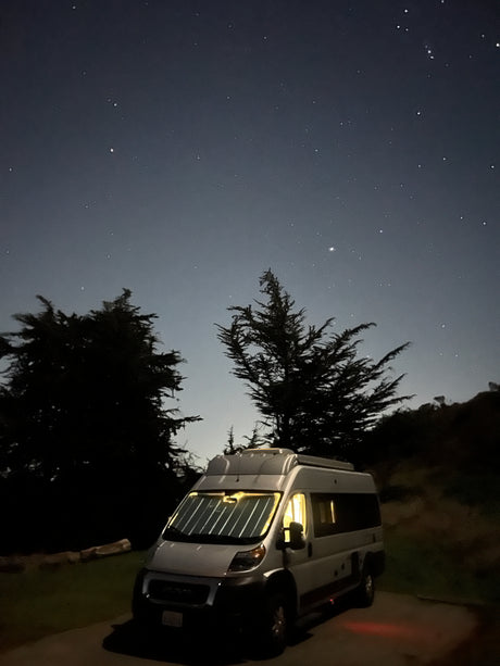 A white camper van, likely an RV rental, illuminates the dark, serene night, parked under a sky full of bright stars, flanked by dark trees.
