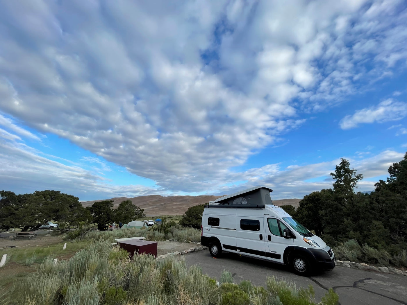 White RV rental camper van at a campsite overlooking sand dunes. Cloudy blue sky above this serene camping scene. — photo 16