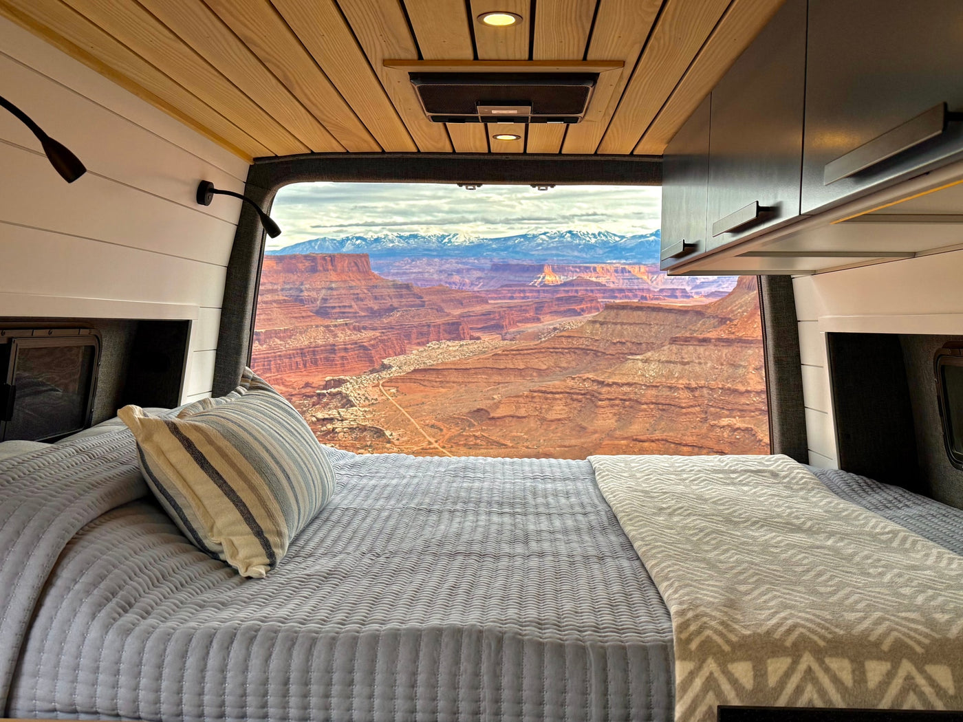 Inside a cozy RV rental, a neatly made bed offers panoramic views of red rock canyons & distant snow-capped mountains. — photo 3