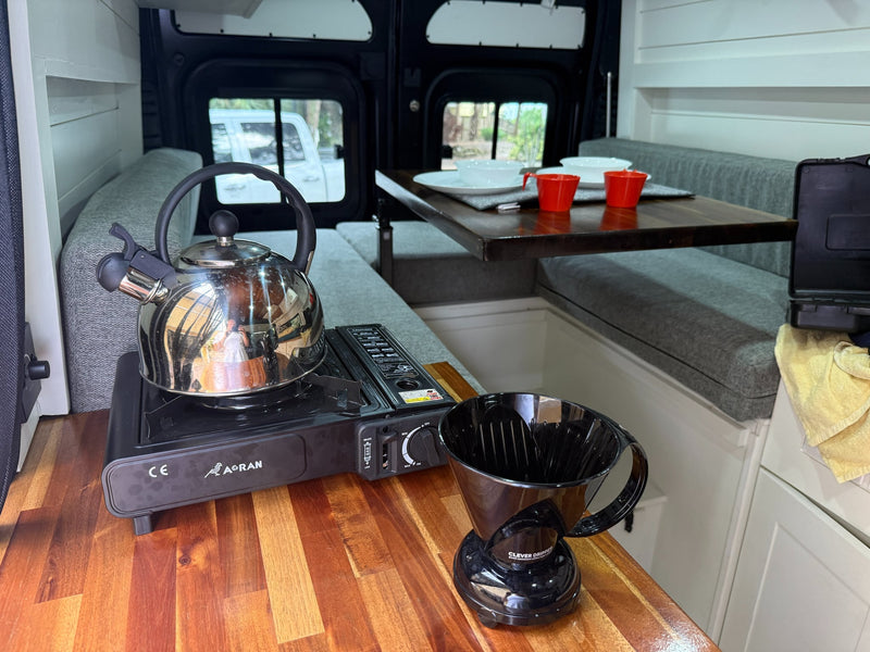  and an American flag. A dog peeks from the mirror.|Cozy camper van interior for your next rv rental adventure. Features a custom dining area with wooden table & comfy benches.|RV rental camp kitchen: portable gas stove — photo 6