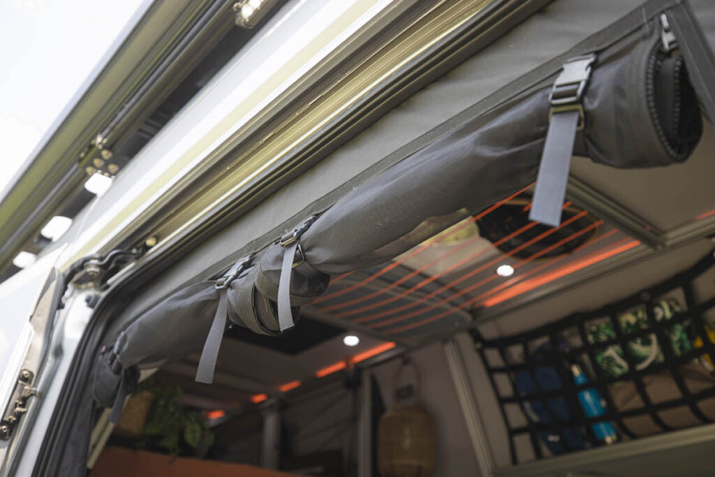 RV rental interior shows a rolled fabric shade/storage, practical overhead lighting, and a gear-filled cargo net, ideal for van life.