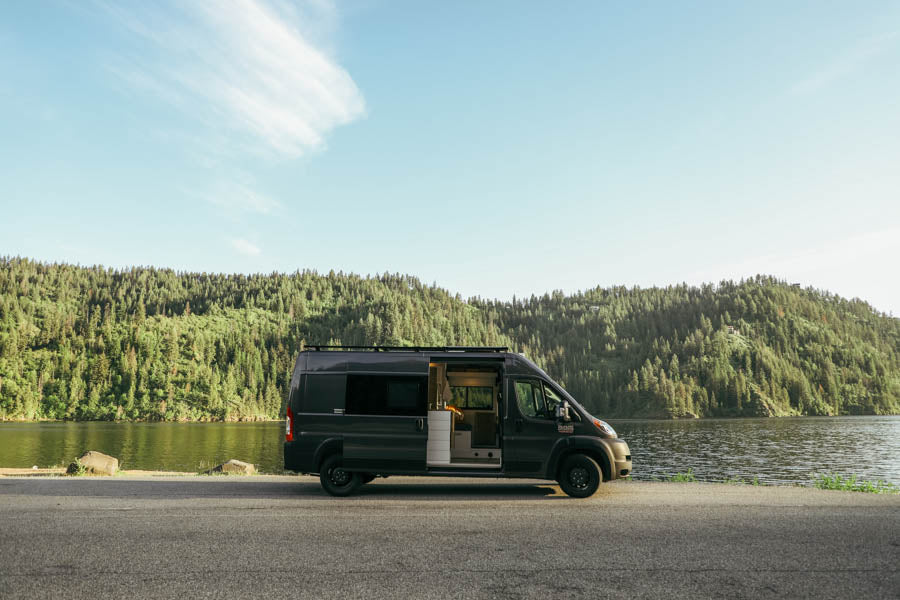 Dark grey RV rental camper van parked by a lake, with green forests & blue sky. Ideal for nature adventures.